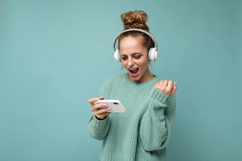 有吸引力的情感积极的年轻的女人穿蓝色的毛衣孤立的蓝色的背景穿白色蓝牙无线耳机听音乐移动电话玩在线游戏gadjet屏幕庆祝赢得