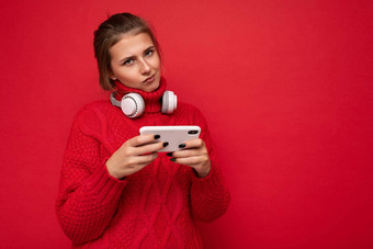 有吸引力的不满意年轻的褐色女人穿红色的毛衣孤立的红色的背景墙持有移动电话冲浪互联网穿蓝牙耳机相机