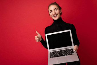<strong>照片</strong>美丽的微笑快乐年轻的浅黑肤色的女人女人持有电脑移动PC空监控屏幕穿黑色的长袖相机给砰砰手势孤立的红色的<strong>墙背景</strong>