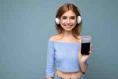 美丽的快乐微笑年轻的女人穿时尚的休闲装孤立的背景墙持有显示移动电话空显示模型穿白色蓝牙耳机听音乐有趣的相机