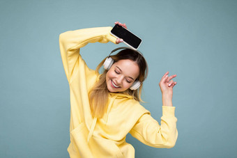 有吸引力的积极的微笑年轻的女人穿时尚的休闲装孤立的色彩鲜艳的背景墙持有显示移动电话空屏幕断路穿白色蓝牙耳机有趣的跳舞