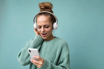 美丽的积极的年轻的女人穿蓝色的毛衣孤立的蓝色的背景穿白色蓝牙无线耳机听音乐移动电话沟通在线互联网gadjet显示