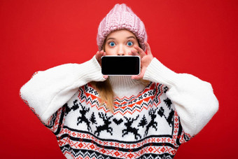 照片有吸引力的惊讶年轻的金发女郎女人穿温<strong>暖</strong>的针织他冬天温<strong>暖</strong>的毛衣站孤立的<strong>红色</strong>的背景显示智能手机空屏幕模拟设备显示
