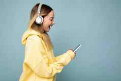 side-ptofile照片美丽的惊讶年轻的女人穿时尚的休闲装孤立的色彩斑斓的背景墙穿白色无线耳机听音乐移动电话消息传递短信智能手机显示