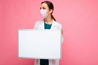 成人有吸引力的女医生保护脸面具白色医疗外套持有空白色磁董事会孤立的粉红色的背景墙