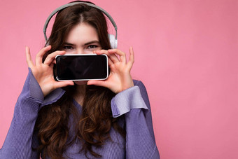 有吸引力的积极的年轻的女人穿时尚的休闲装孤立的色彩鲜艳的背景墙持有显示移动电话空屏幕断路穿白色蓝牙耳机有趣的相机