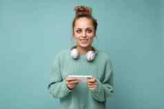 有吸引力的积极的年轻的女人穿蓝色的毛衣孤立的蓝色的背景穿白色蓝牙无线耳机听音乐移动电话沟通在线互联网相机