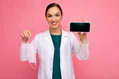 有吸引力的微笑医疗年轻的女人穿白色外套持有面具显示现代移动电话孤立的粉红色的背景相机