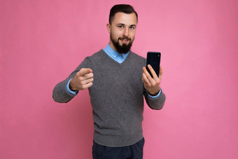 好快乐很酷的年轻的浅黑肤色的女人不剃须的男人。beardwearing时尚的灰色毛衣蓝色的衬衫站孤立的粉红色的背景墙持有智能<strong>手机电</strong>话相机指出<strong>手</strong>指相机