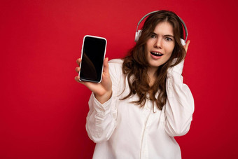 有吸引力的积极的震惊了年轻的浅黑肤色的女人卷曲的女人穿白色衬衫孤立的红色的背景墙持有显示移动电话空显示穿白色蓝牙耳机听音乐相机