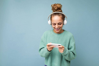 美丽的积极的年轻的女人穿蓝色的毛衣孤立的蓝色的背景穿白色蓝牙无线耳机听音乐移动电话沟通在线互联网gadjet显示