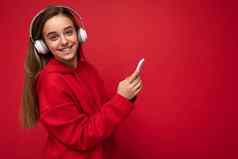 侧面轮廓拍摄积极的微笑漂亮的浅黑肤色的女人女孩穿红色的连帽衫孤立的红色的背景持有智能手机写作短信消息穿白色无线耳机听很酷的音乐相机