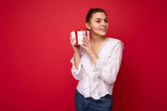 拍摄有吸引力的积极的微笑年轻的浅黑肤色的女人女人孤立的红色的背景墙穿白色上衣持有白色礼物盒子红色的丝带一边