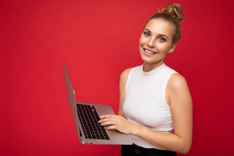 一边配置<strong>文件</strong>照片拍摄美丽的微笑金发碧眼的年轻的女人聚集头发穿白色t恤持有电脑移动PC打字<strong>键盘</strong>相机孤立的红色的墙背景