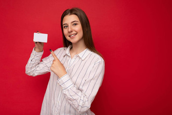照片拍摄有吸引力的积极的微笑年轻的黑暗金发女郎女人穿白色上衣孤立的红色的背景持有信贷卡相机指出手指塑料非接触式卡