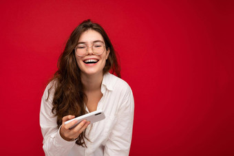 照片漂亮的微笑积极的年轻的浅黑肤色的女人女人穿白色衬衫光学眼镜孤立的红色的背景持有手移动电话玩在线游戏相机有趣的