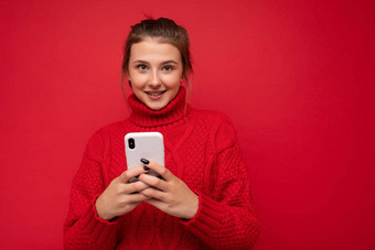 有吸引力的可爱的微笑年轻的女人穿温暖的<strong>红色</strong>的毛衣孤立的<strong>红色</strong>的<strong>背景墙</strong>持有智能手机沟通在线互联网相机
