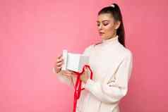 有吸引力的快乐惊讶年轻的浅黑肤色的女人女人孤立的粉红色的背景墙穿白色毛衣持有礼物盒子unboxing现在相机