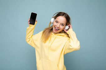 照片美丽的快乐的微笑年轻的女人穿时尚的休闲衣服孤立的背景墙持有移动电话穿白色蓝牙耳机听音乐有趣的相机