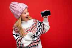 照片有吸引力的微笑年轻的金发女郎女人穿温暖的针织他冬天温暖的毛衣站孤立的红色的背景显示智能手机空屏幕模拟设备显示