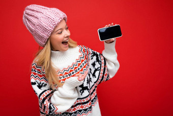 照片有吸引力的微笑年轻的金发女郎女人穿温暖的针织他冬天温暖的毛衣站孤立的红色的背景显示智能<strong>手机</strong>空屏幕<strong>模拟</strong>设备显示