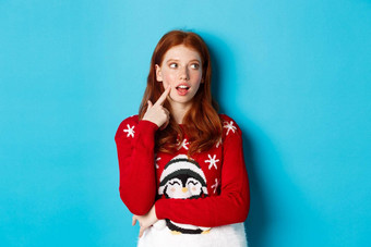 冬天假期圣诞节夏娃概念漂亮的红色头发的人女孩圣诞节毛衣触碰脸颊深思熟虑的微笑使选择上角落里思考蓝色的背景