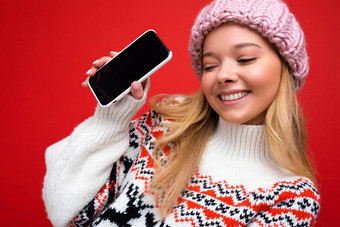 照片有吸引力的微笑年轻的金发女郎女人穿温暖的针织他冬天温暖的毛衣站孤立的红色的背景显示智能手机空屏幕模拟