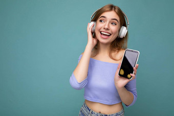 美丽的快乐微笑年轻的女人穿时尚的休闲装孤立的背景墙持有显示移动电话空显示模型穿白色<strong>蓝牙耳机</strong>听音乐有趣的一边