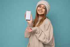 照片美丽的微笑积极的好年轻的女人穿时尚的米色毛衣米色针织冬天他平衡孤立的蓝色的背景空空间持有手显示移动电话空屏幕模型相机