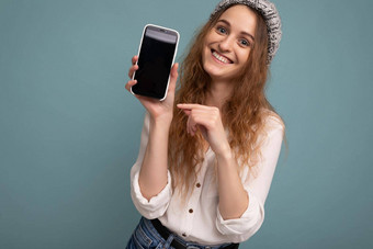 特写镜头照片美丽的积极的年轻的金发女郎卷曲的女人穿休闲白色衬衫灰色他孤立的蓝色的背景墙持有电话显示智能手机空dcreen模型相机