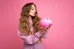 肖像怀疑思考年轻的美丽的有吸引力的女人金发女郎头发真诚的情绪穿时尚的粉红色的连帽衫孤立的粉红色的背景复制空间持有粉红色的小猪盒子Moneybox概念