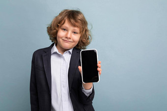 快乐微笑英俊的积极的男孩金发碧眼的卷曲的头发穿西装持有电话孤立的蓝色的背景相机显示智能手机空显示屏幕