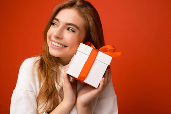 特写镜头肖像照片美丽的快乐金发女郎年轻的女人孤立的橙色背景墙穿白色连帽衫持有白色礼物盒子红色的丝带一边享受