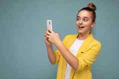 美丽的年轻的惊讶女孩穿黄色的夹克白色t恤站孤立的蓝色的背景冲浪互联网电话移动屏幕