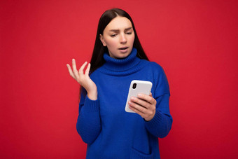 心烦意乱有吸引力的积极的好年轻的浅黑肤色的女人女人穿时尚的蓝色的温暖的毛衣平衡孤立的红色的背景空空间持有手移动电话消息传递短信设备理解