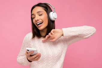照片美丽的快乐的微笑年轻的女人穿时尚的休闲衣服孤立的背景墙持有移动电话穿白色蓝牙耳机听音乐有趣的一边
