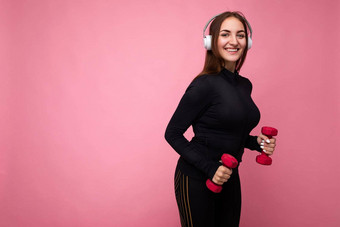 有吸引力的积极的微笑年轻的浅黑肤色的女人女人穿黑色的体育运动衣服孤立的粉红色的背景墙健身哑铃穿白色蓝牙耳机听好音乐有趣的相机