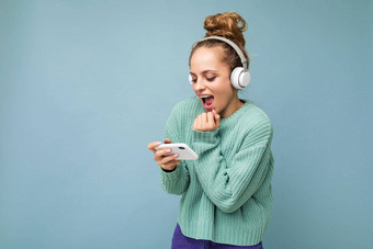 有吸引力的情感积极的年轻的女人穿蓝色的毛衣孤立的蓝色的背景穿白色蓝牙无线耳机听音乐移动电话玩在线游戏gadjet屏幕有关