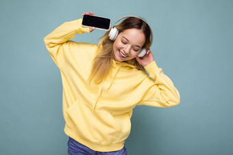 美丽的快乐微笑年轻的女人穿时尚的休闲装孤立的背景墙持有显示移动电话空显示模型穿白色<strong>蓝牙耳机</strong>听音乐有趣的