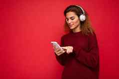 拍摄美丽的年轻的浅黑肤色的女人卷曲的女人穿黑暗红色的毛衣孤立的红色的背景墙穿白色无线耳机听音乐移动电话冲浪互联网智能手机显示