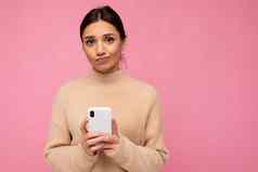 有吸引力的可爱的不开心年轻的褐色女人穿米色温暖的毛衣站孤立的粉红色的背景冲浪互联网电话相机