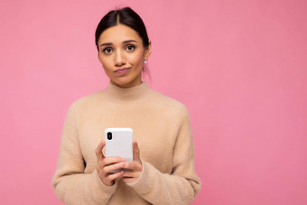 有吸引力的可爱的不开心年轻的褐色女人穿米色温暖的毛衣站孤立的粉红色的背景冲浪互联网电话相机