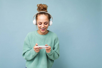 美丽的积极的微笑年轻的女人穿蓝色的毛衣孤立的蓝色的背景穿白色蓝牙无线耳机听音乐移动电话沟通在线互联网gadjet显示