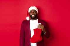 英俊的非洲美国男人。庆祝冬天假期持有圣诞节袜子微笑穿圣诞老人他站红色的背景
