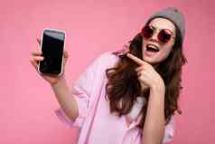 照片积极的有吸引力的年轻的浅黑肤色的女人女人穿时尚的粉红色的衬衫灰色他色彩鲜艳的太阳镜孤立的粉红色的背景持有手显示移动电话空显示模型相机指出手指设备