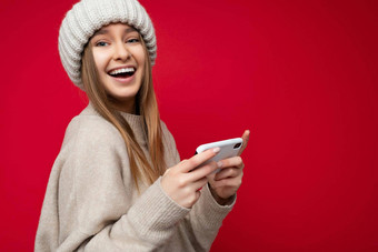 美丽的快乐的快乐年轻的金发女郎女人穿休闲米色毛衣米色他孤立的红色的背景持有手移动电话沟通在线互联网相机