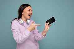 照片美丽的积极的笑年轻的褐色女人穿紫色的连帽衫孤立的蓝色的背景持有智能手机穿白色无线耳机一边指出手指电话