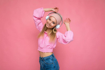 照片有吸引力的积极的微笑年轻的金发女郎女人穿粉红色的上衣粉红色的他孤立的粉红色的背景墙穿白色无线蓝牙耳机听音乐有趣的相机