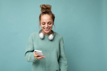有吸引力的笑积极的年轻的女人穿蓝色的毛衣孤立的蓝色的背景穿白色蓝牙无线耳机听音乐移动电话沟通在线互联网