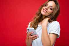 漂亮的可爱的年轻的金发女郎女人穿白色t恤站孤立的红色的背景冲浪互联网电话相机